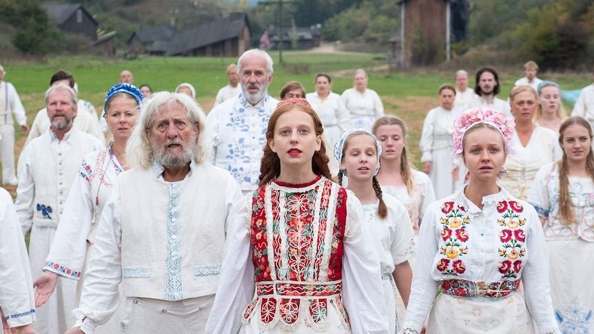 A group of people in white clothes covered in colourful flowers stare mournfully into the distance.
