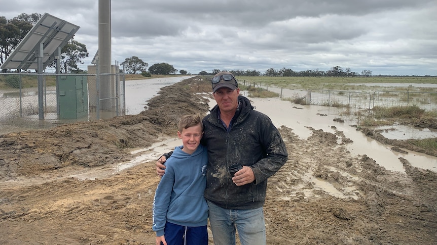 A man with one arm around a boy in front of a flooded paddock.