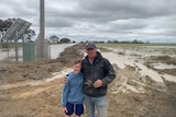 A man with one arm around a boy in front of a flooded paddock.