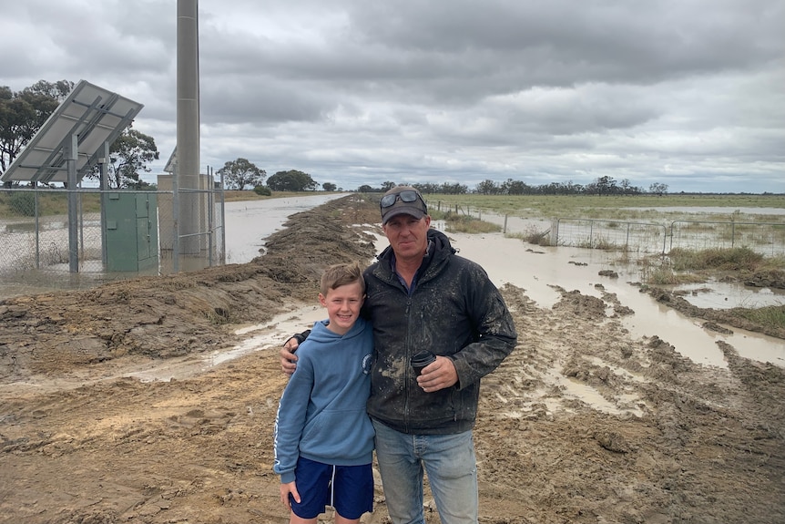 A man with one arm around a boy in front of a flooded paddock.