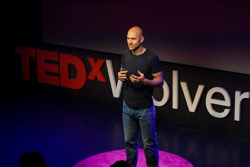 A young man with shaved hair, wears black tee, blue jeans, stands on stage talking. Red and white Tedxvolver sign behind him.
