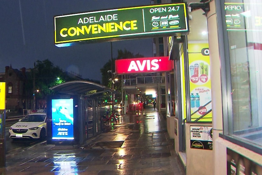 A convenience store with a police car outside at a bus stop in the rain at night