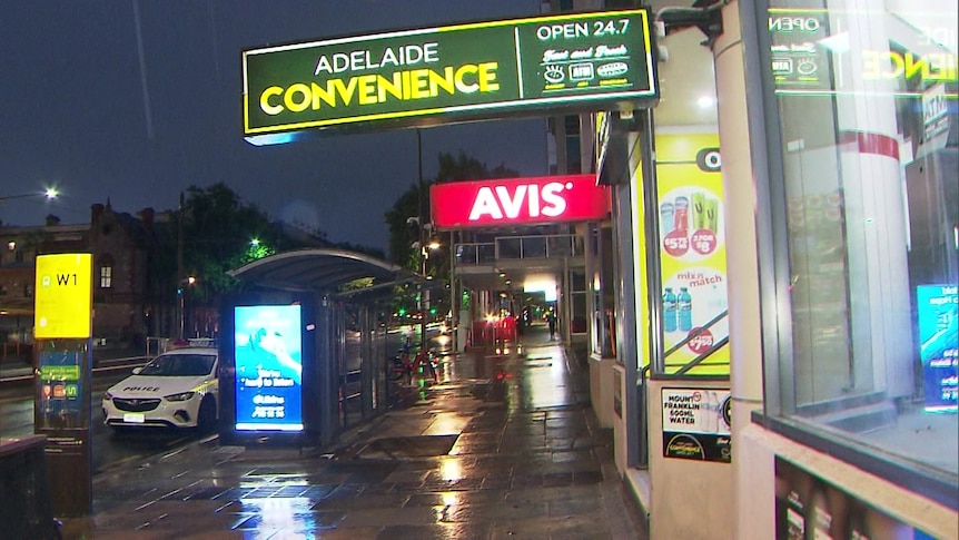 A convenience store with a police car outside at a bus stop in the rain at night