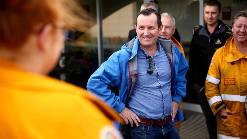 Mark McGowan in wet weather gear stands hands on hips smiling at volunteers in fire fighting uniforms.