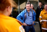 Mark McGowan in wet weather gear stands hands on hips smiling at volunteers in fire fighting uniforms.
