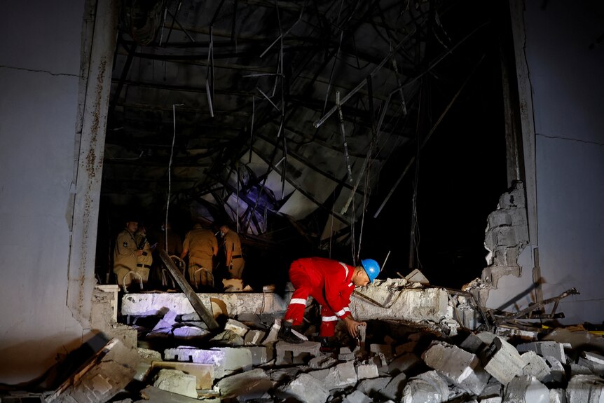 A man in an orange emergency uniform picks up a brick on the top of a pile of rubble.