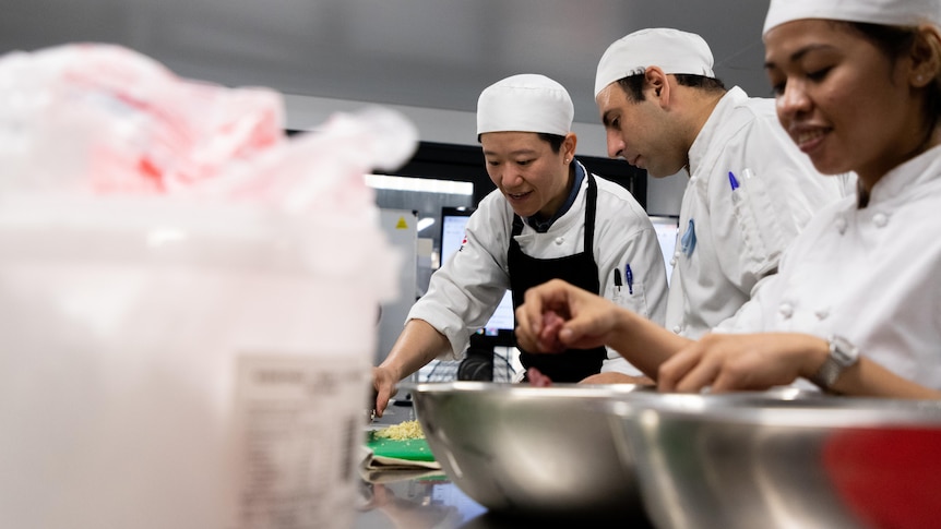 Three people in chef uniforms working on a stainless steel bench with ingredients.