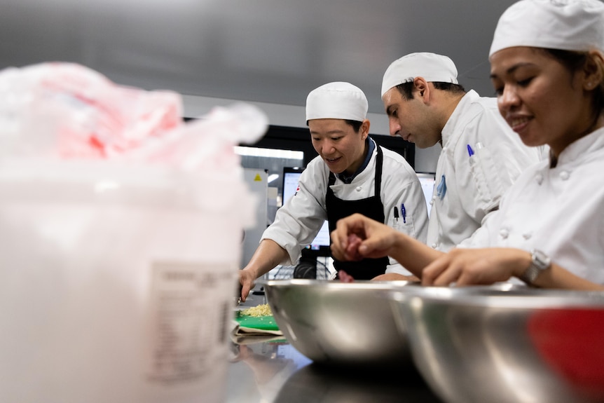 Three people in chef uniforms working on a stainless steel bench with ingredients.
