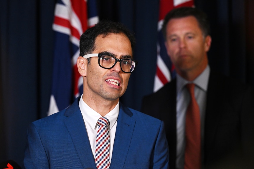a man wearing glasses speaking at a press conference while another man stands behind him looking