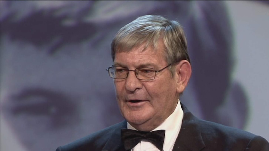 AFL great Royce Hart speaks at a podium dressed in a suit.