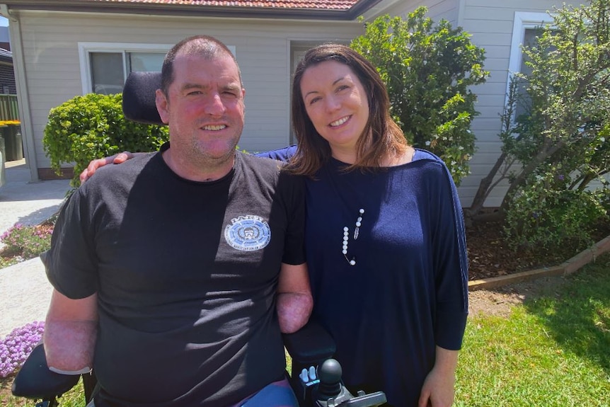 A man in a wheelchair, with no limbs, smiles next to his wife who has brown hair. They stand outside their home