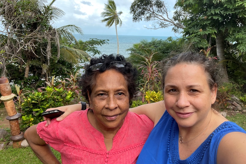 Mother and daughter stand together in front of palm trees and the ocean.