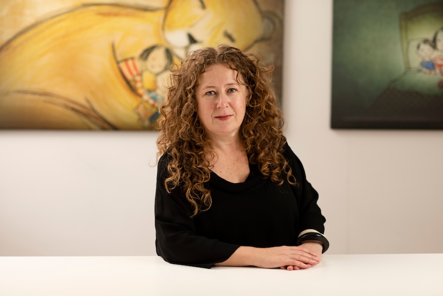 Michal Morris, a woman with long, brown curly hair, rests her forearms on a white table