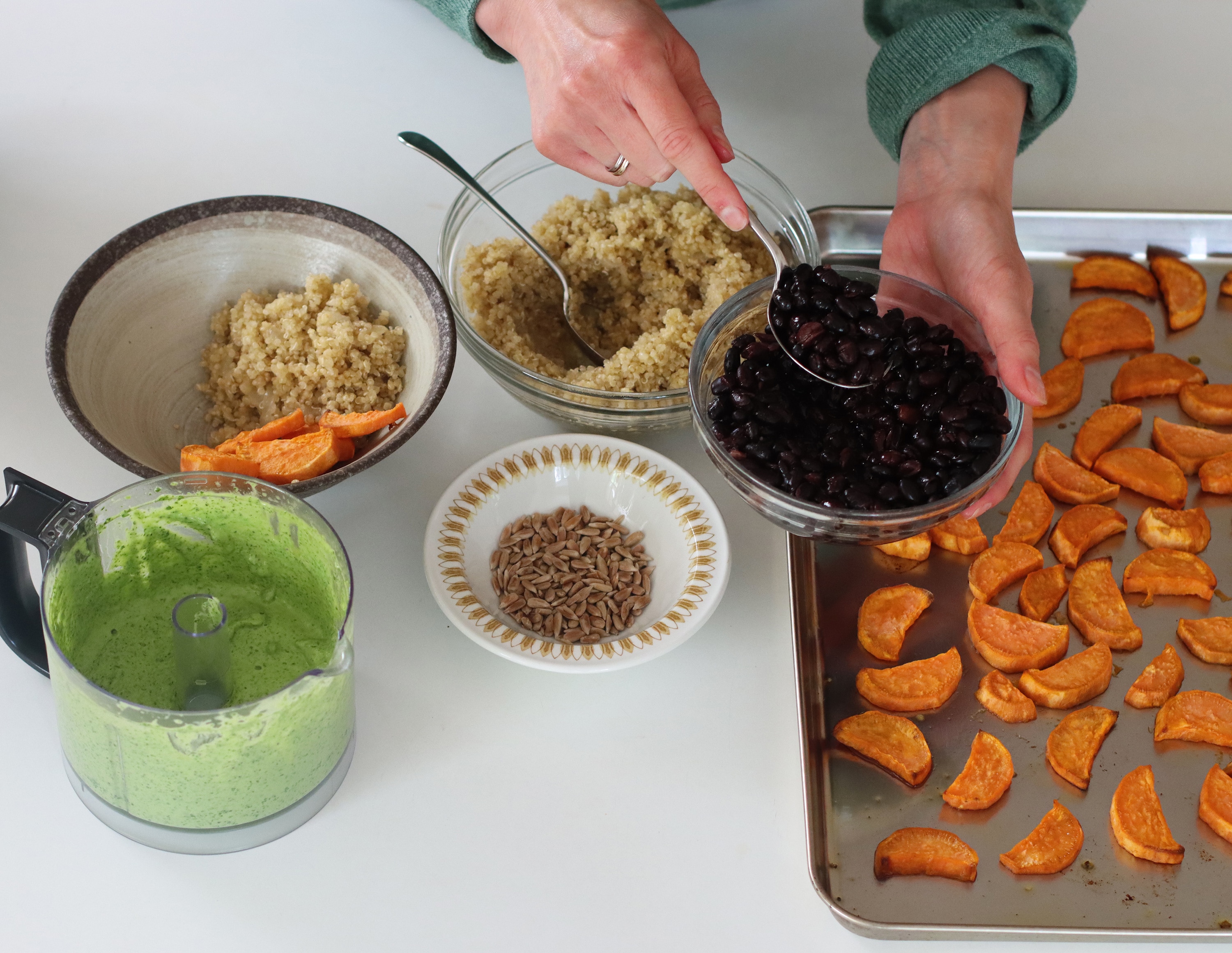 Close-up of hands scooping black beans to be added to a bowl with quinoa, roasted sweet potato and green tahini sauce.