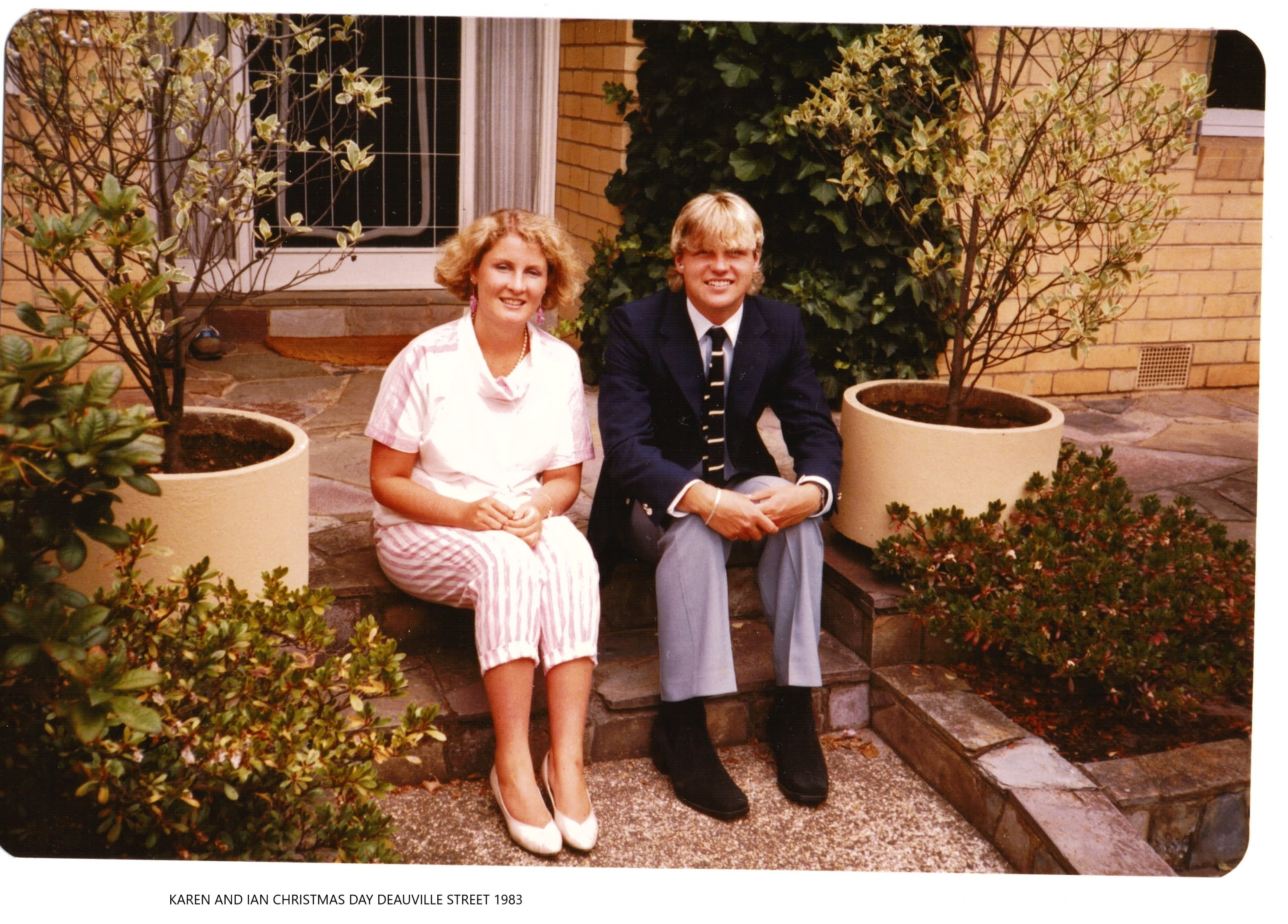 A young woman and man, both blonde, sit together on a doorstep of a house, with large pot plants either side of them.