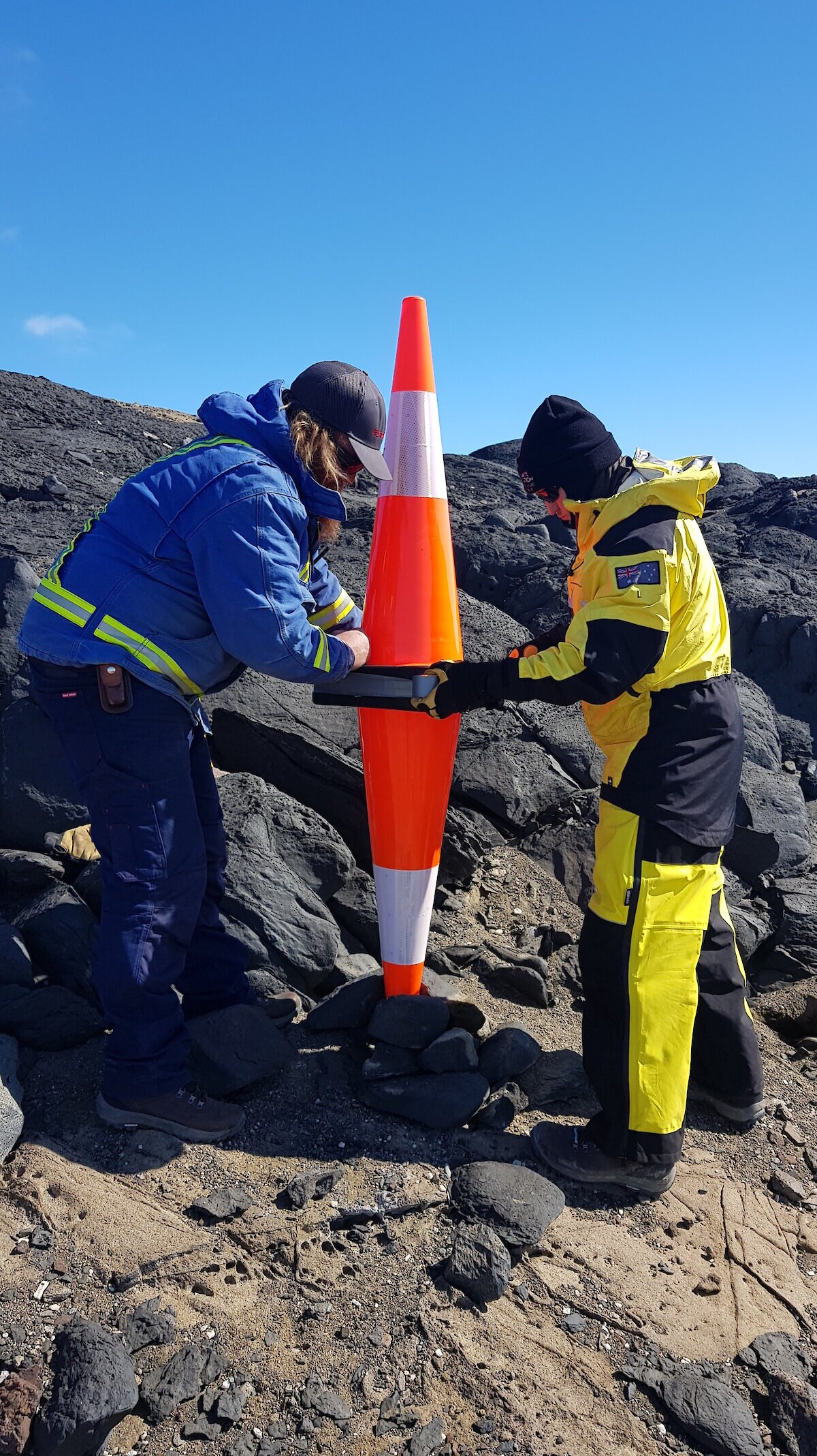 Two Antarctic expeditioners either side of two traffic cones on rocky ground