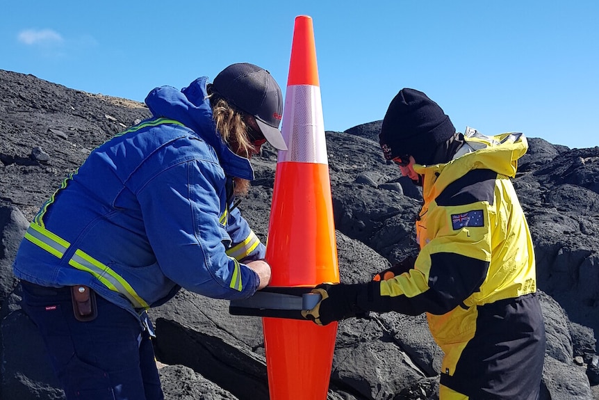 Two Antarctic expeditioners either side of two traffic cones on rocky ground