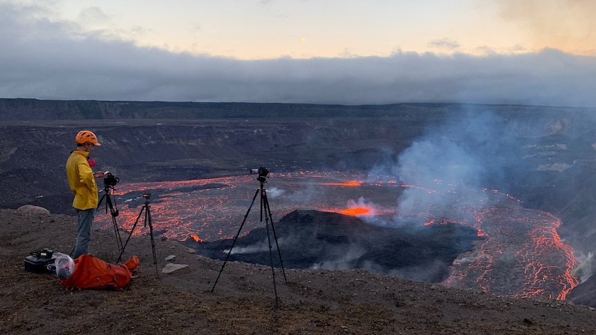 A person stands at volcano rim with camera gear on tripod.