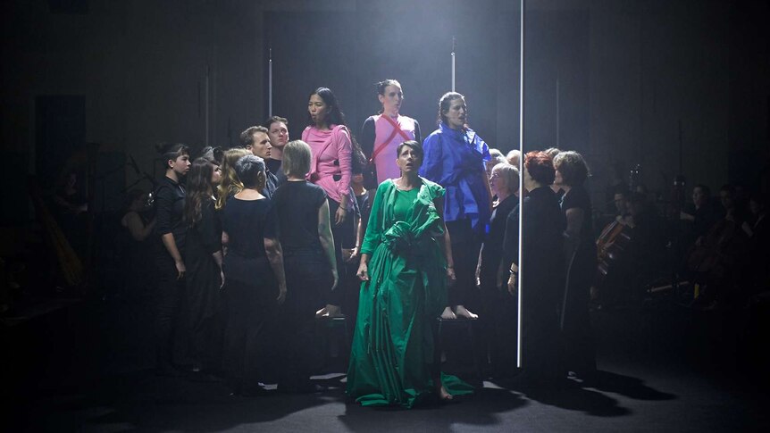 Cluster of performers centre stage, darkened backdrop, haze above the group, four soloists in colourful clothes sing at centre.