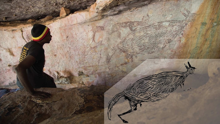 An Indigenous man sits in front of a rock wall on which kangaroos are painted