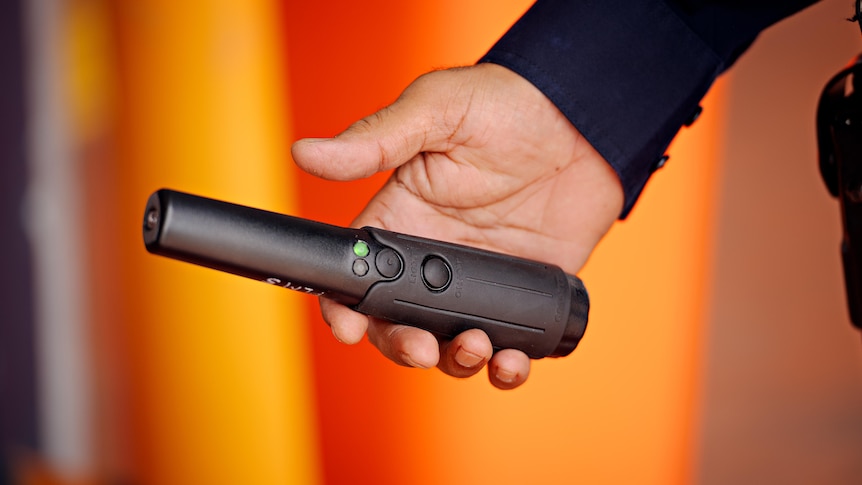 A person holds a black wanding device with a green light on it