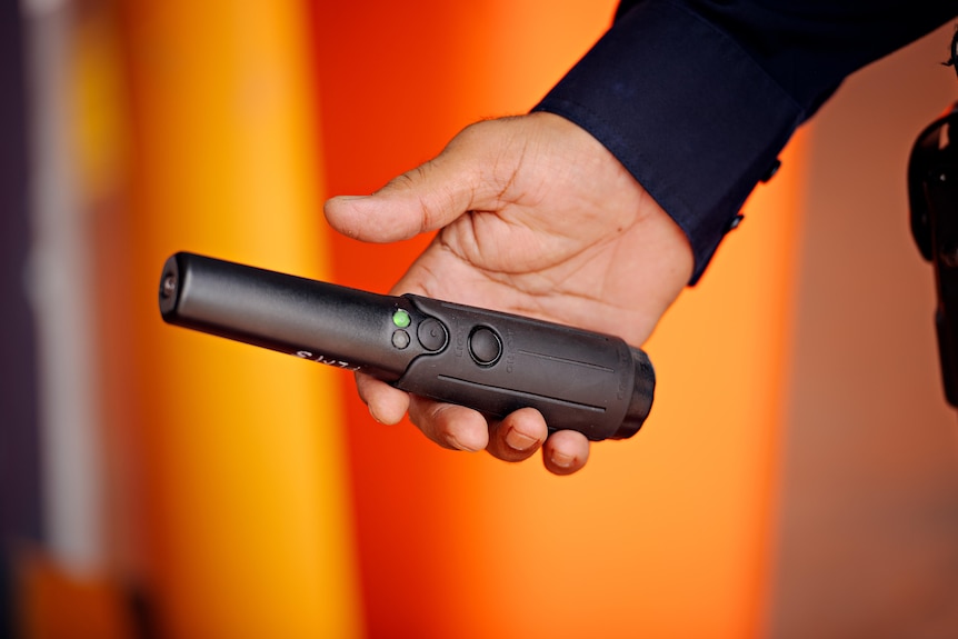 A person holds a black wanding device with a green light on it