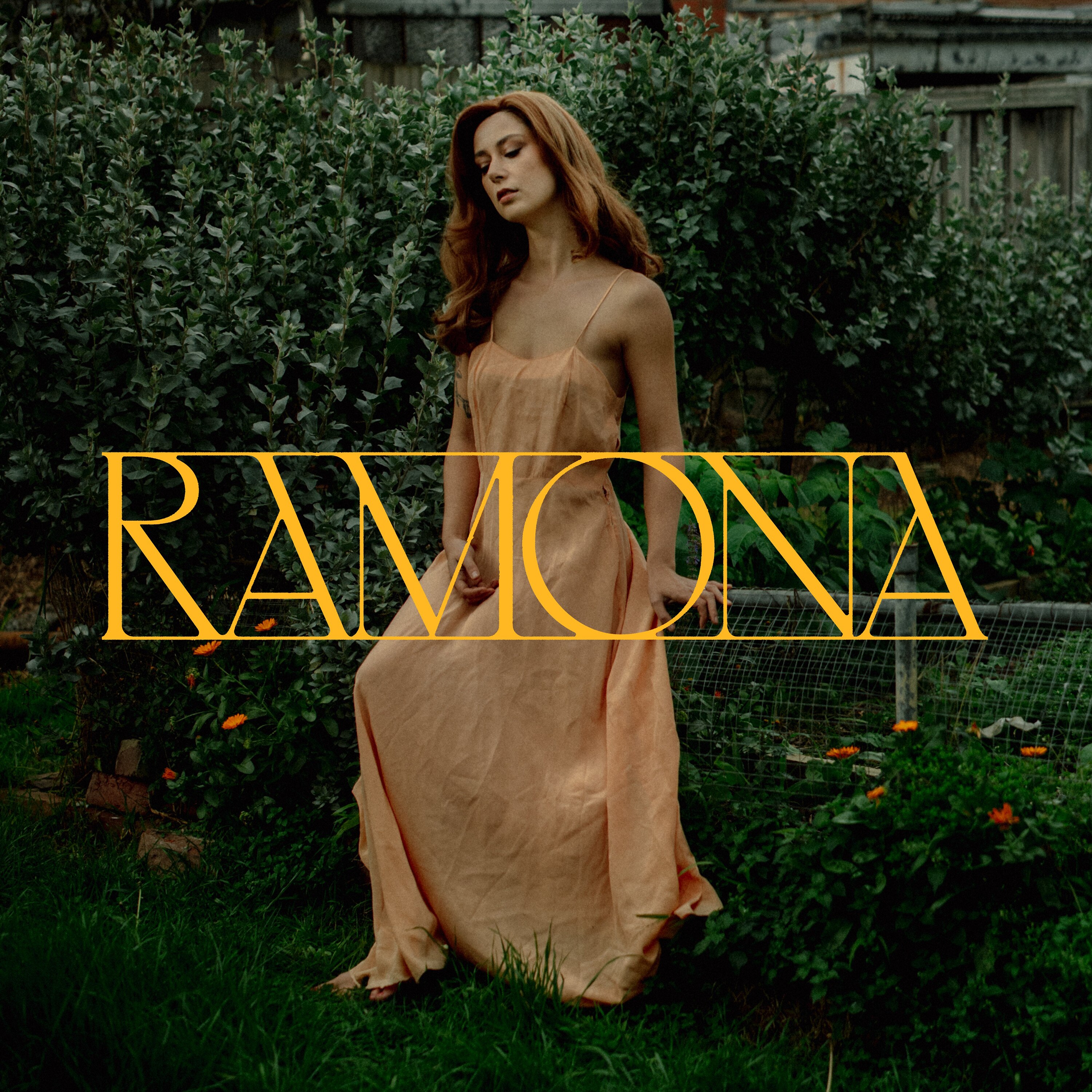 Brunette woman stands in tan cloth-gown by hedge & low-fenced garden fence with text: RAMONA