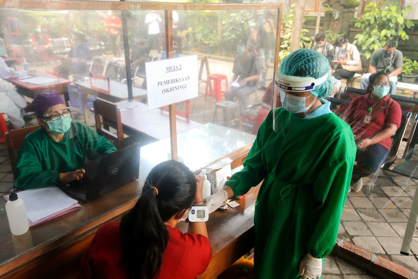 A person wearing a mask sits behind a shield as a woman administers a health check on a woman sitting down.