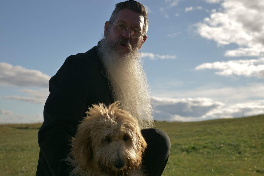 Former Safari Park Keeper Ron Prendergast holding a dog, in a green expanse, in front of a blue sky, near Gisborne.