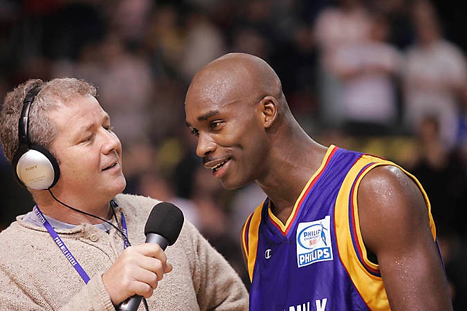 A man wearing headphones holds up a microphone to interview a smiling basketball player