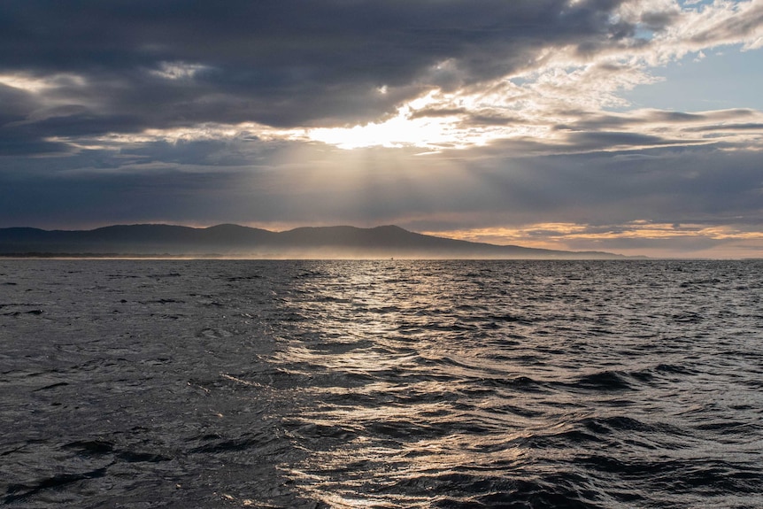 A shot over the ocean, dark clouds on horizon with the sun peaking through, hills on horizon, a boat far away on water.