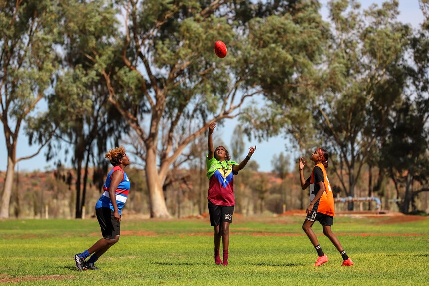 Two young Aboriginal women wearing football jumpers prepare to jump at a football thrown by an umpire