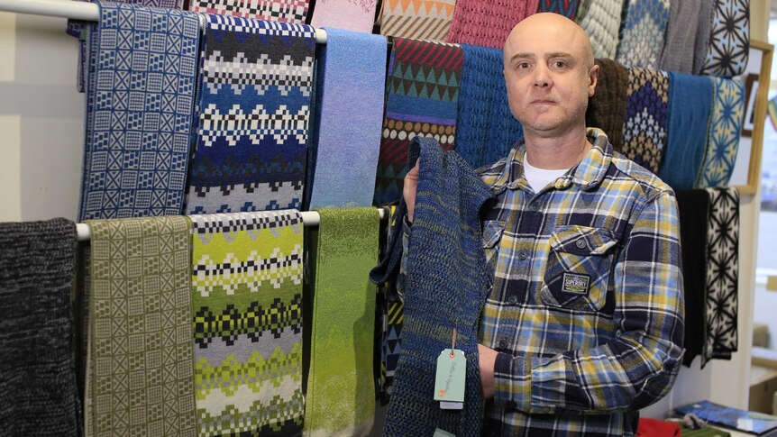 Anthony Mananov holds up a blue scarf while standing in front of several racks of different coloured scarves.