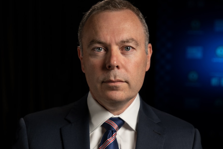 Raptor squad commander Jason Weinstein wears a suit and stares directly ahead, against a dark backdrop.