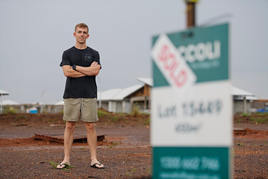 A 24-year-old man stands with his arms folded, smiling on his newly purchased block of land. A sold sign is at the forefront. 