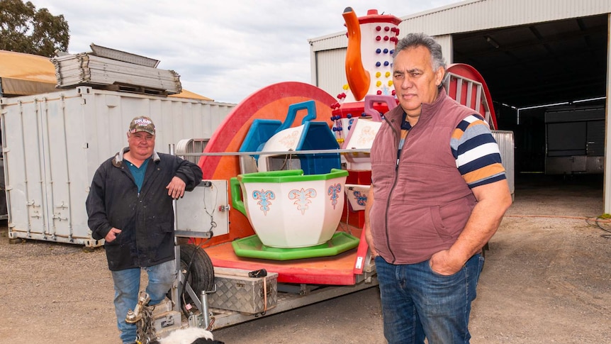 Two men and a dog in front of an amusement ride on a trailer.