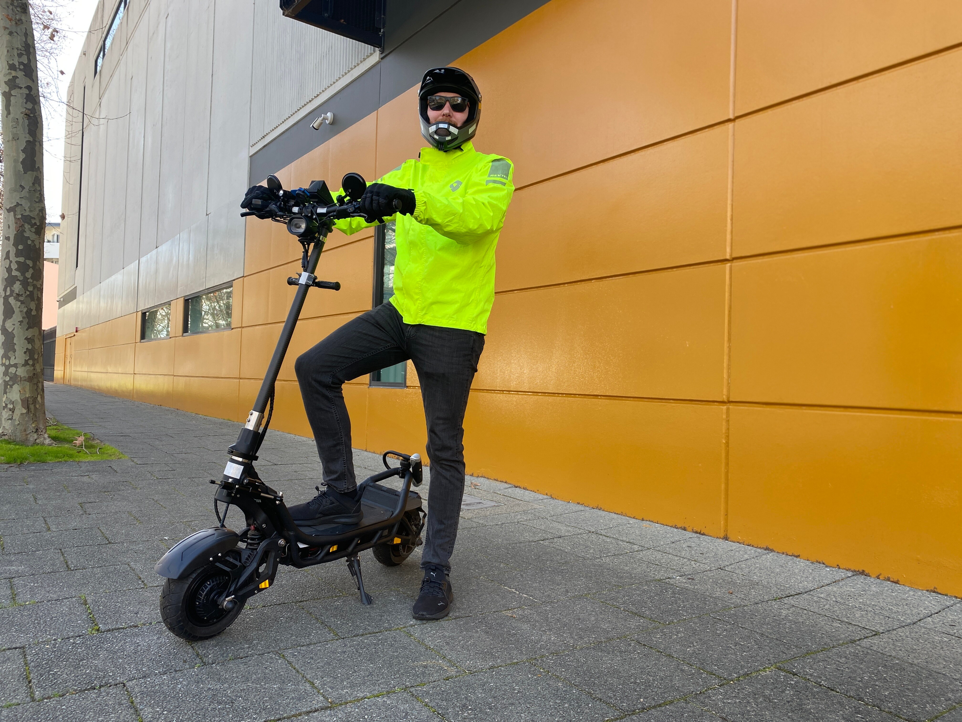 A man in high-vis and a helmet on an e-scooter on a city street.