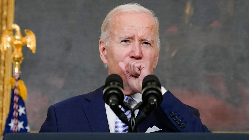 Joe Biden covers his mouth with a fist as he stands in front of microphones at a lectern. 