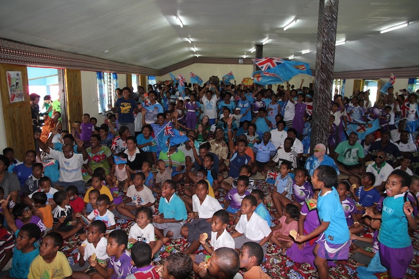 Large group of adults and children gathered in hall, waving flags, cheering and looking away from camera at television