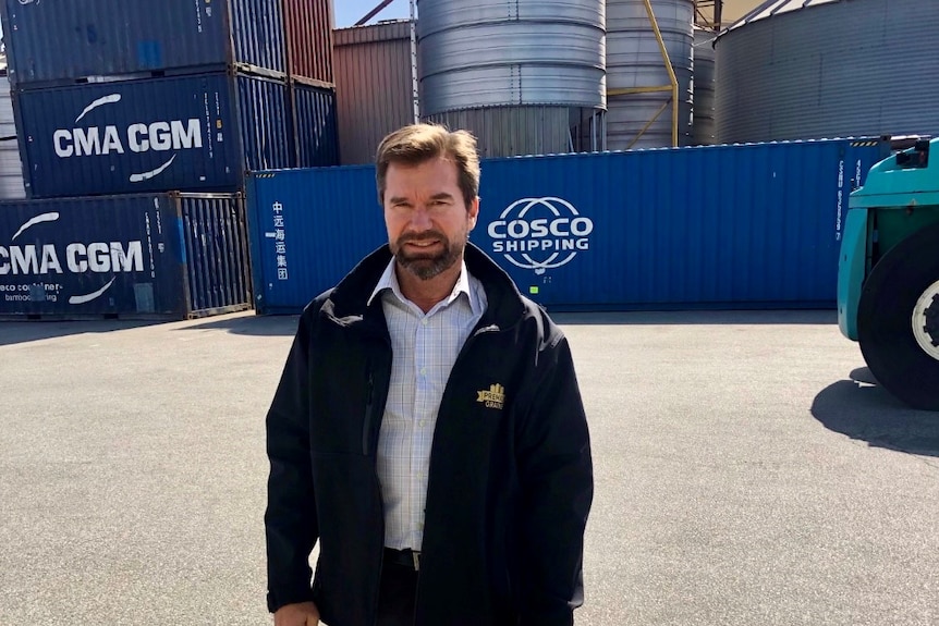 A man with a beard looks at the camera with shipping containers in the background