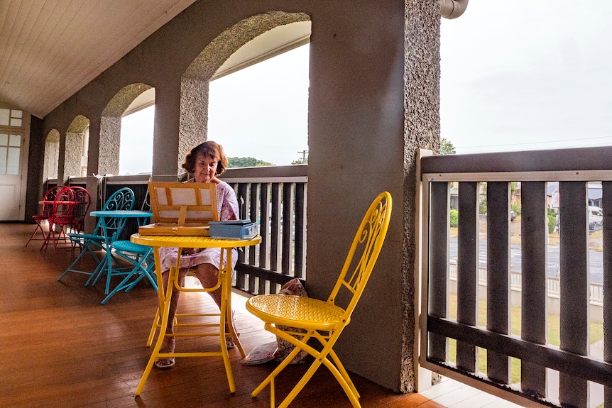 A woman sits working on an easel on a verandah.