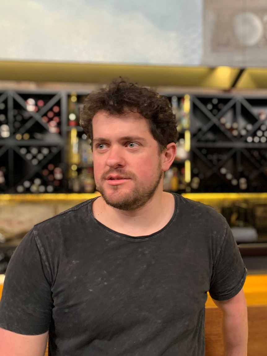 A dark-haired, bearded man in a black shirt stands in front of a bar.