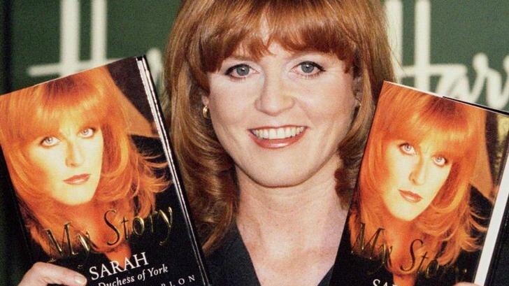 Sarah Ferguson grins at the camera, wearing a black blazer with her hair down as she holds two copies of her book.