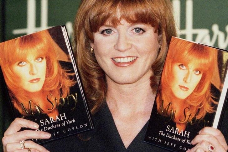 Sarah Ferguson grins at the camera, wearing a black blazer with her hair down as she holds two copies of her book.