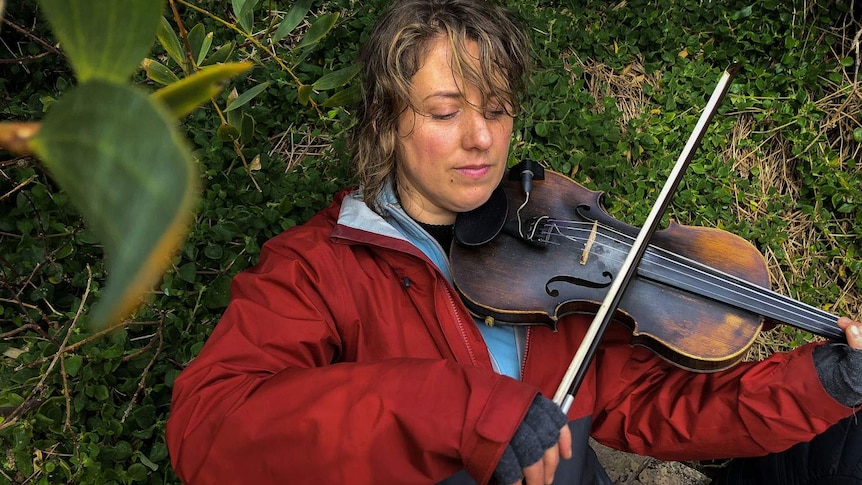 Rachel Meyers playing violin, sheltered by the leafy river bank