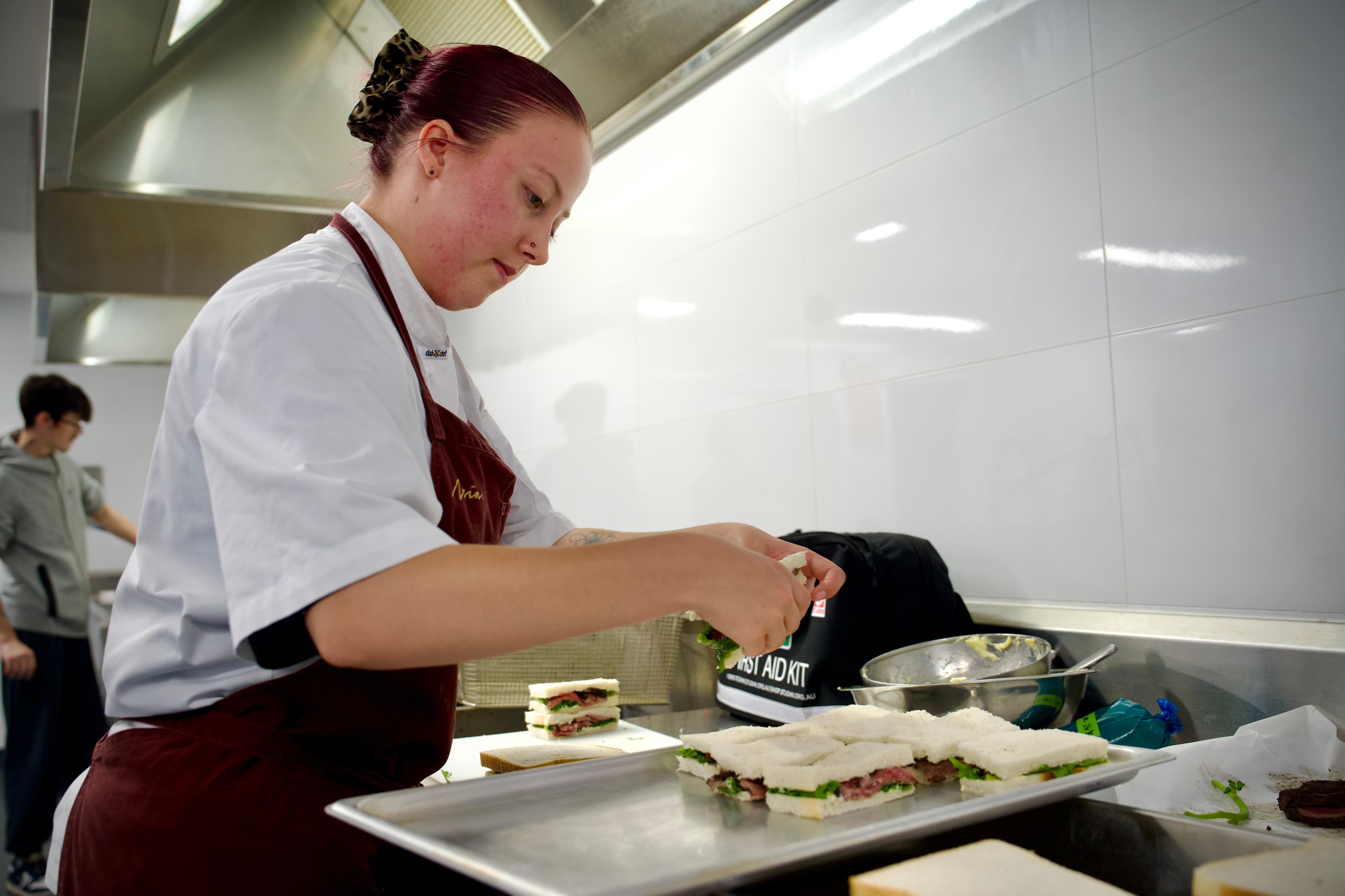 A chefs apprentice wearing chefs whites and an apron stands in a commercial kitchen plating up a dish of food