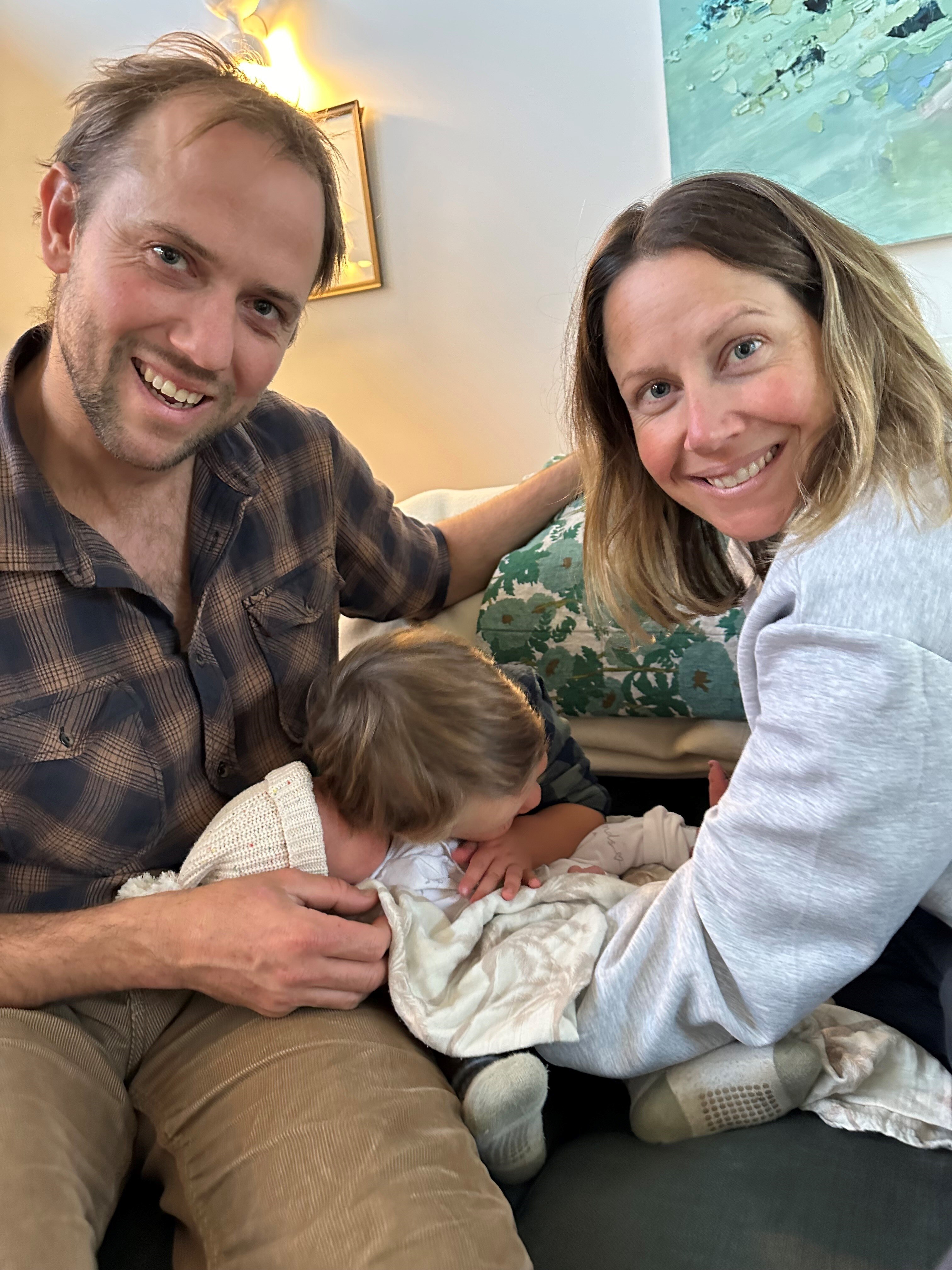 A man in a shirt and brown pants poses with woman in a blue jumper, a young child, and an infant child.