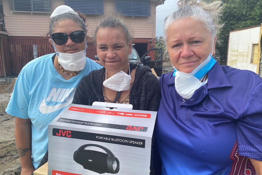 Three women stand in front of a house, with masks around their necks.