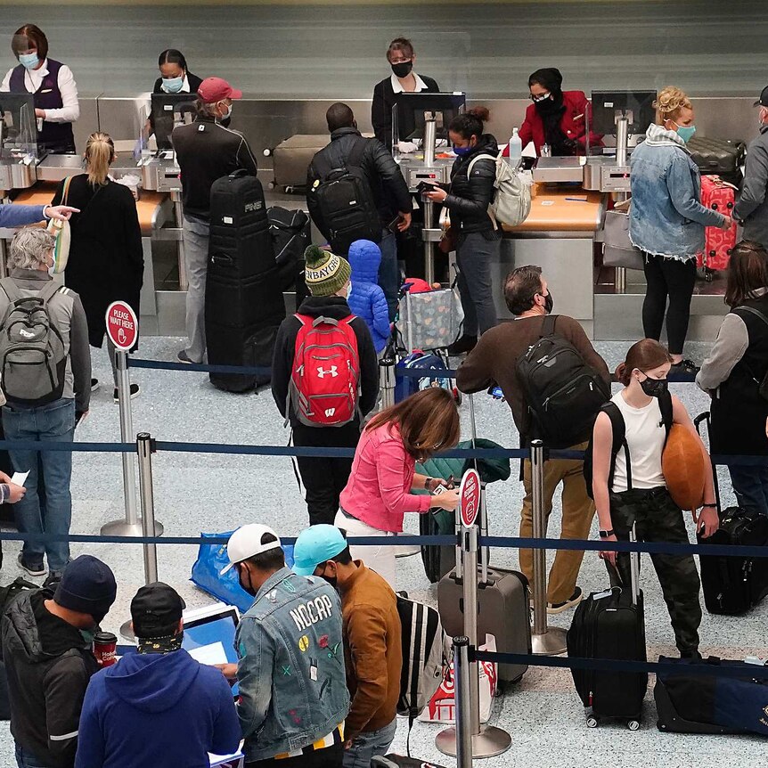 A group of people with their luggage bags stand in line at an airport wearing masks.