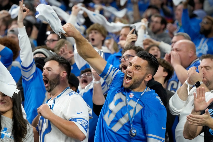 A group of Detroit Lions NFL fans roar, punch the air and wave white towels during a playoff game.